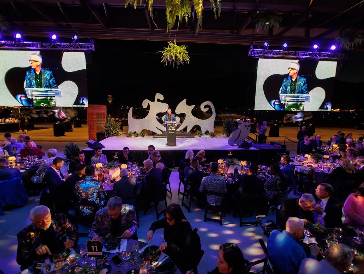 Wide view of a speaker on stage with projection screens and seated guests