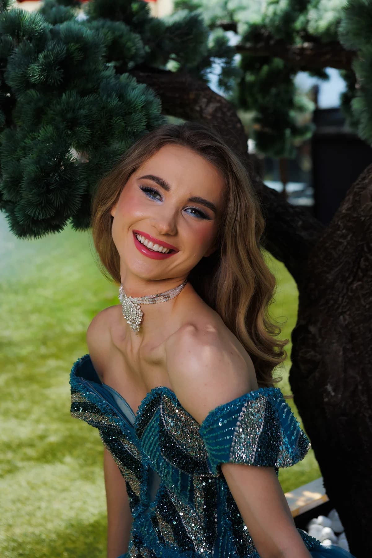 Model smiling in a blue embellished gown beneath a tree