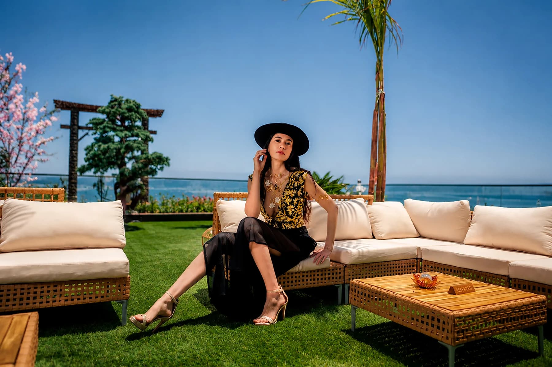 Model in a black hat and patterned top seated on a sofa beneath a palm tree