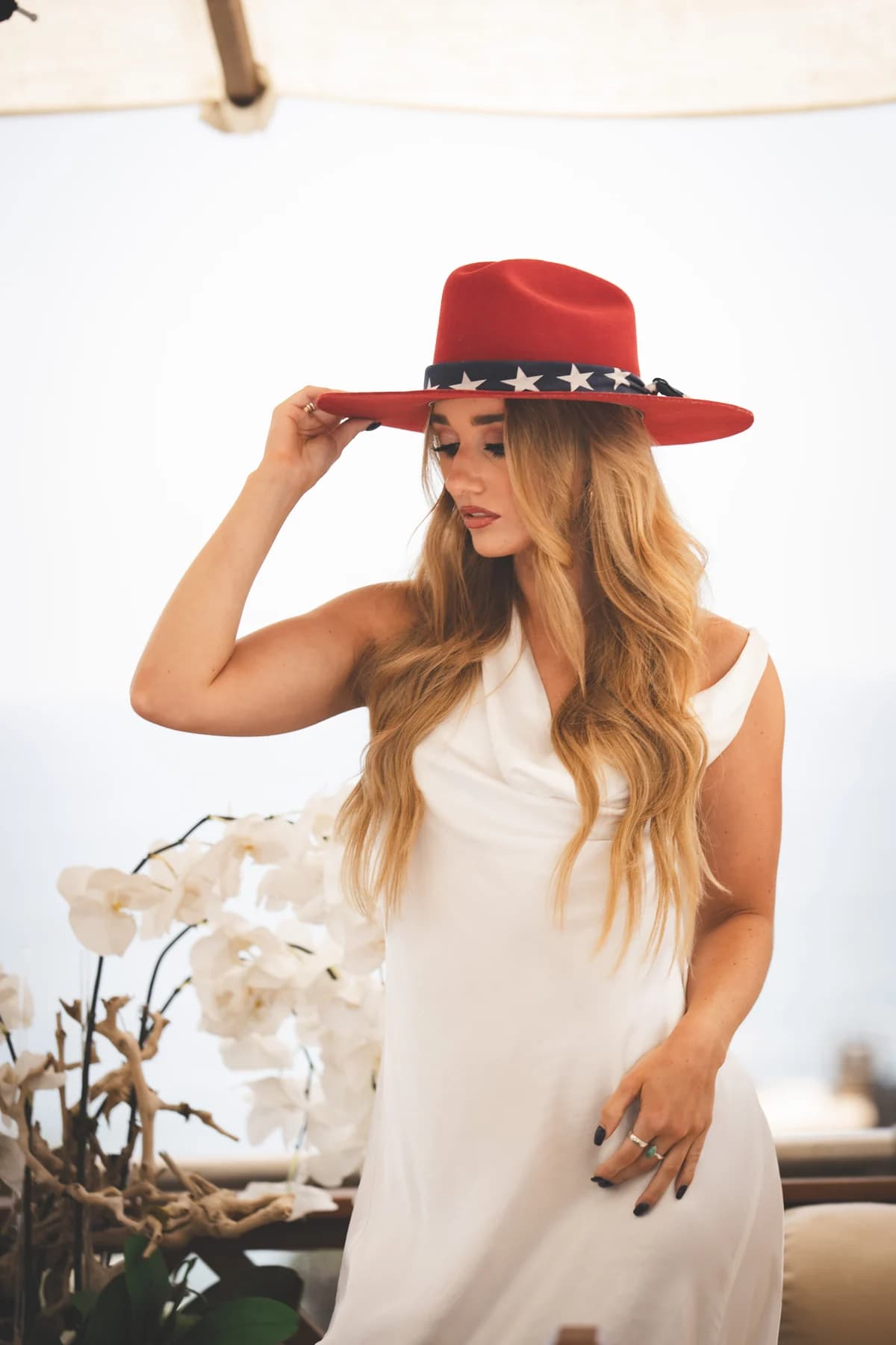 Model in a white outfit holding the brim of a red hat beside white flowers