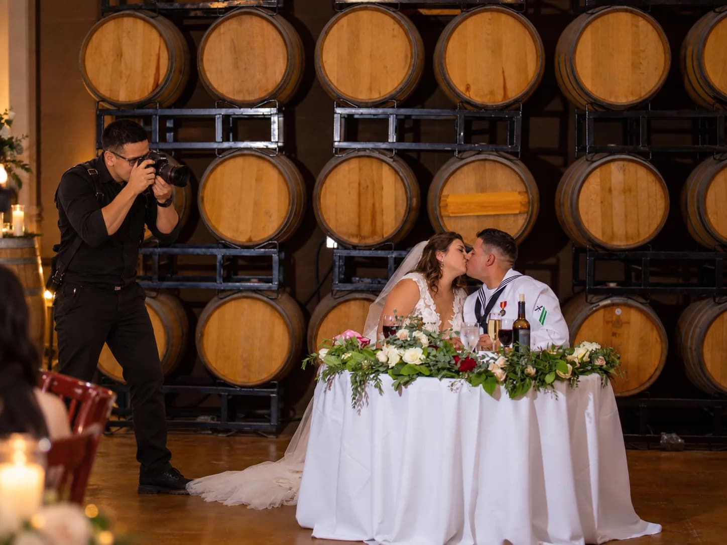 Mario holding a camera beside a wedding couple seated in front of wine barrels