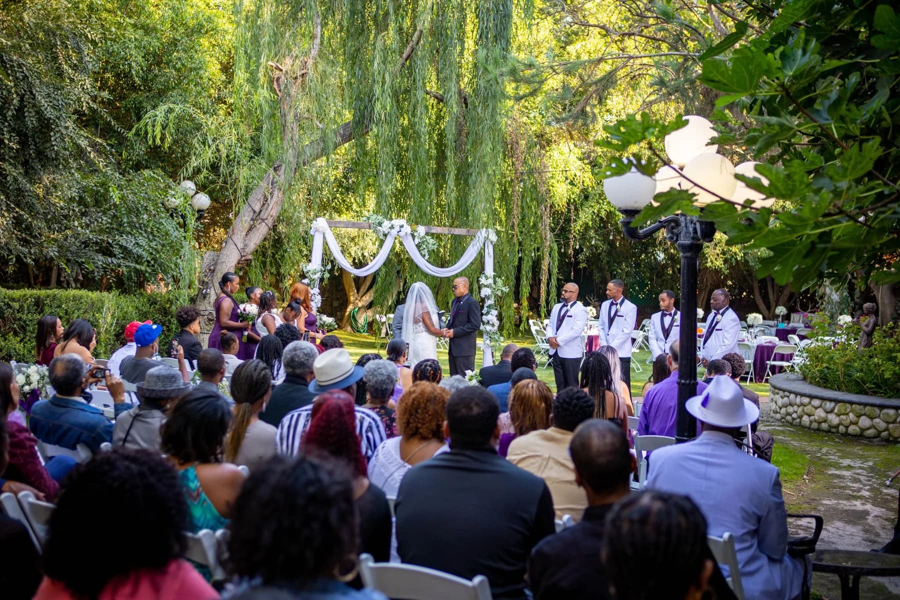 Guests seated beneath trees facing a white-draped garden ceremony arbor