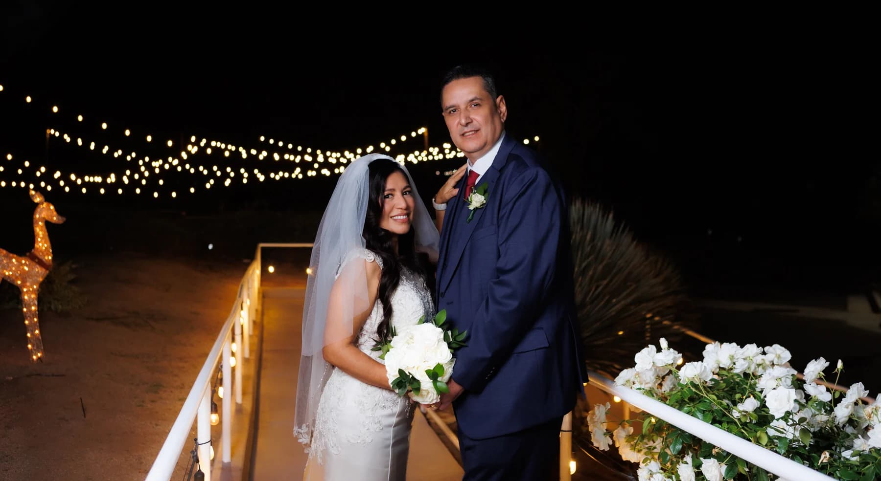Bertha and Xavier holding a white bouquet beside a railing beneath evening string lights
