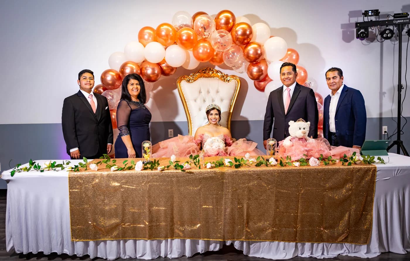 Quinceañera seated with family members beneath a rose-gold balloon arch