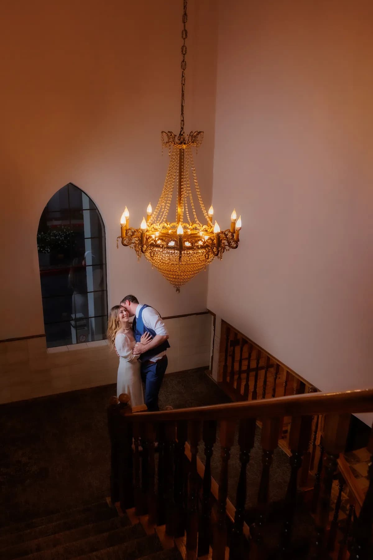 Gloria and Ryan embracing beneath a chandelier beside an arched window