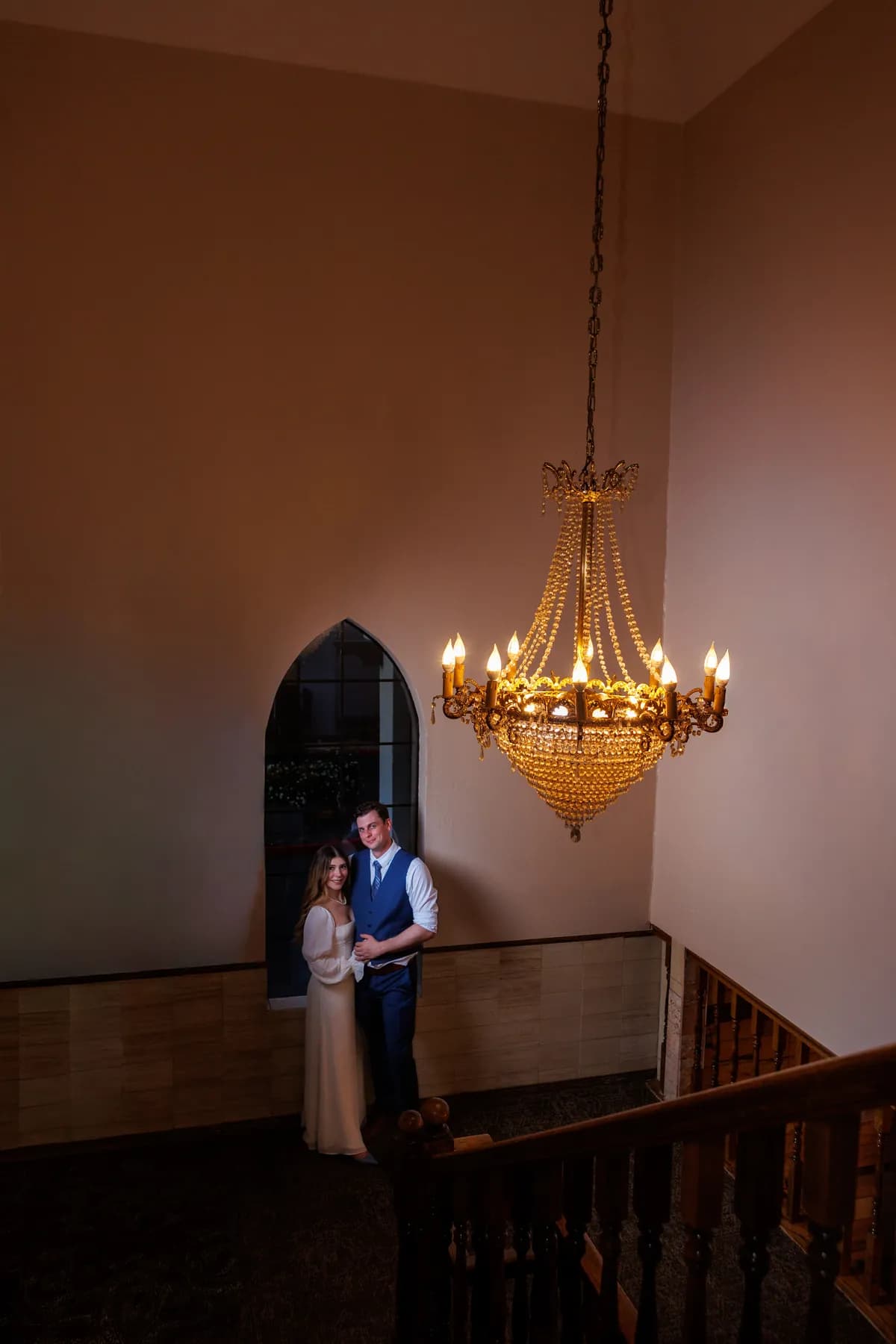 Gloria and Ryan standing close near an arched window and chandelier