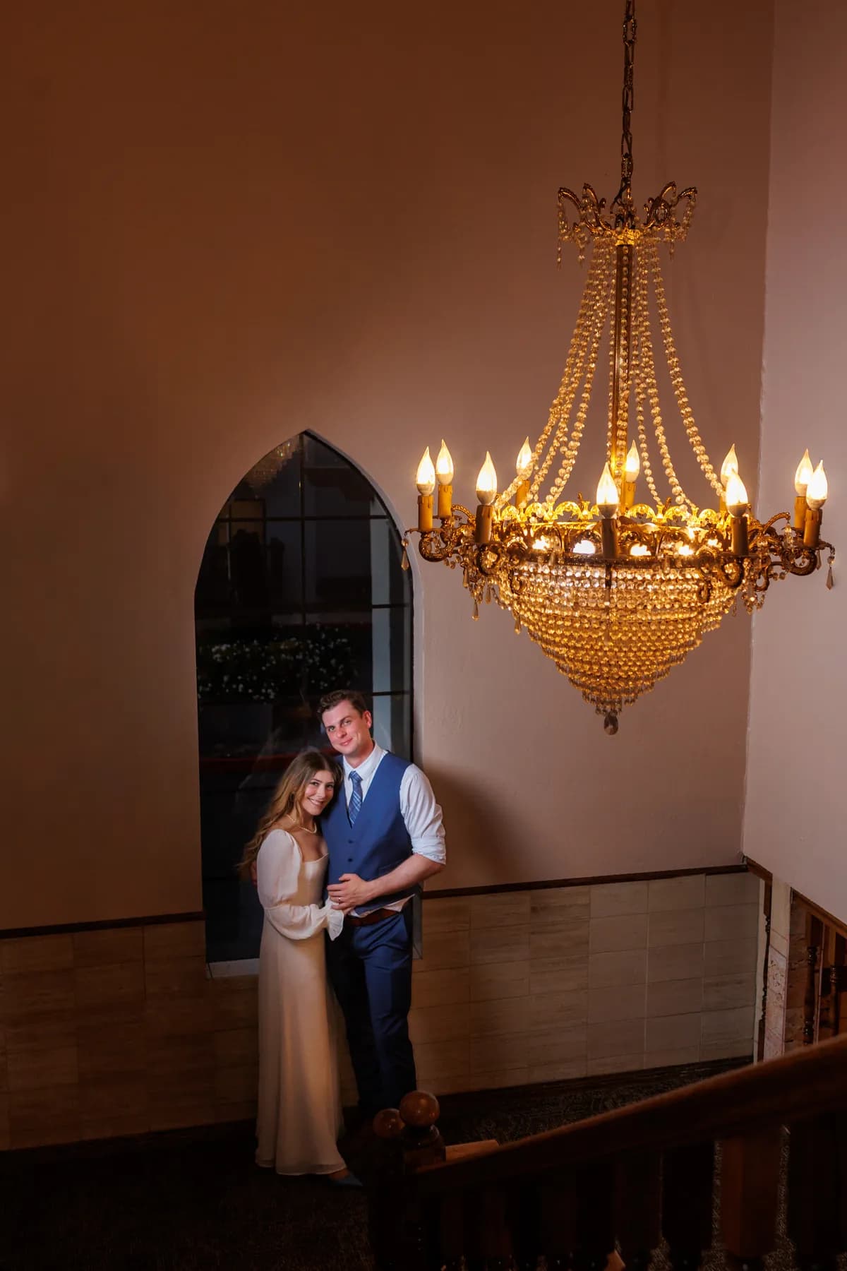 Gloria and Ryan posing beneath a lit chandelier near an arched window