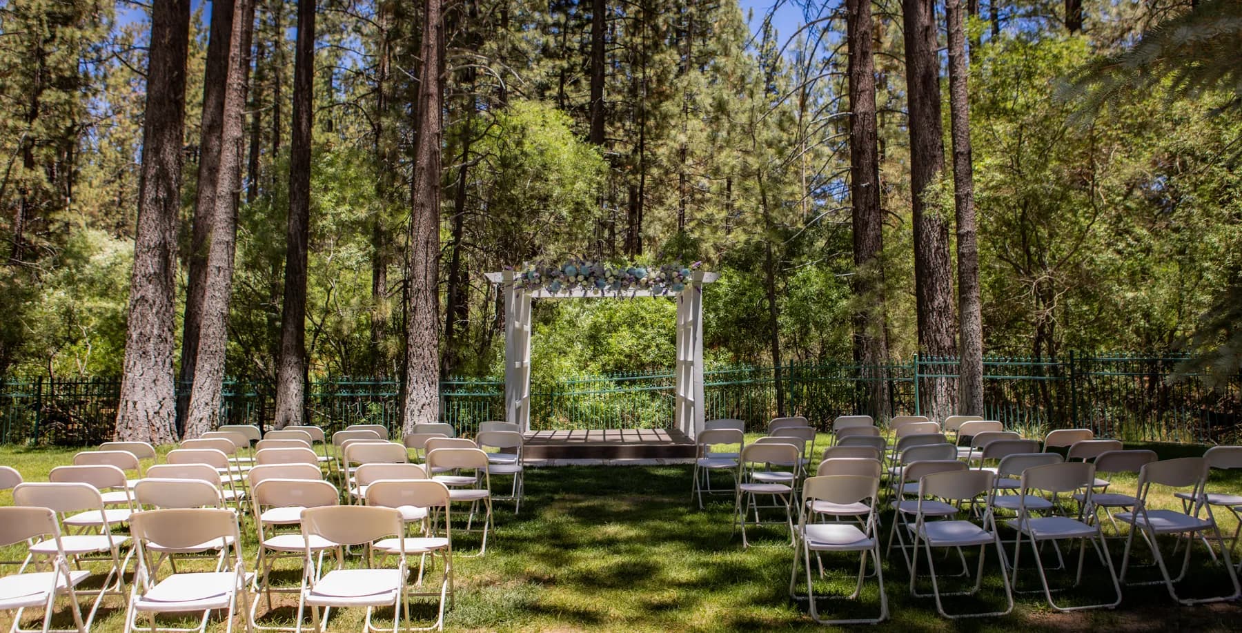 Empty outdoor ceremony chairs facing a floral arbor among tall pines