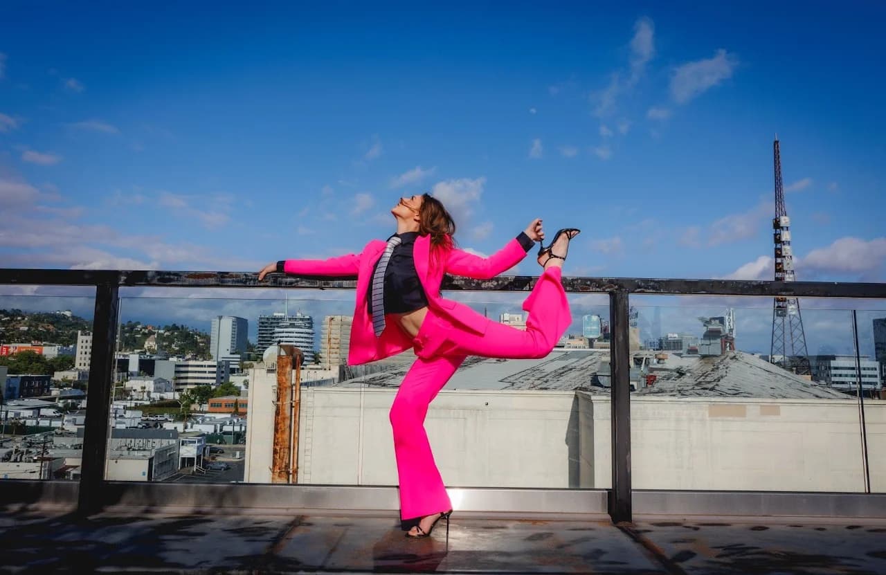 Model in a bright pink suit lifting one leg beside a rooftop railing